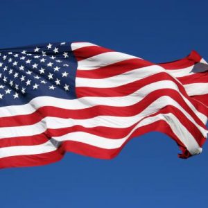 An American flag waving in the wind as it hangs upon a tall flagpole against a solid blue background.