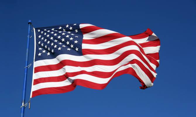An American flag waving in the wind as it hangs upon a tall flagpole against a solid blue background.