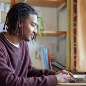 A young college student studying in his dorm room with a laptop in front of him and a window and plants in the background.