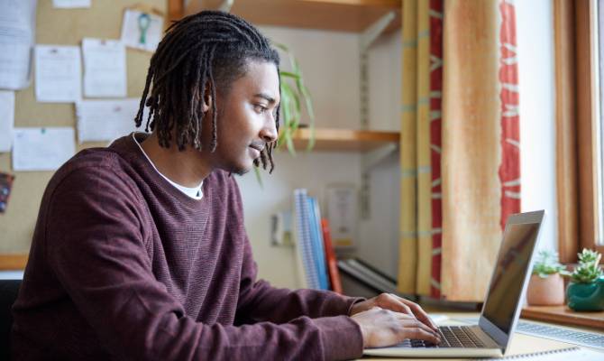 A young college student studying in his dorm room with a laptop in front of him and a window and plants in the background.