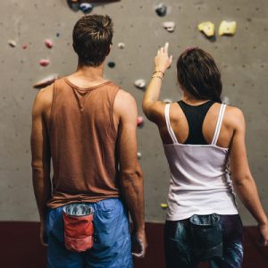 A young man and woman with their back to the viewer looking at an indoor rock climbing wall as the woman points at the wall.