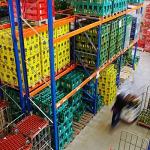 Tall metal storage racks holding colorful crates of beverages in a warehouse. A worker is pushing a cart down an aisle.