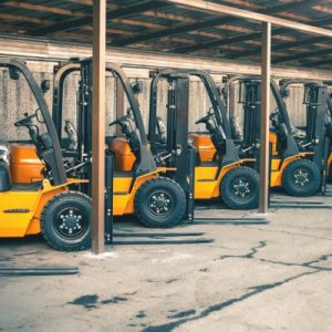 Eight yellow forklifts in like-new condition parked diagonally under a tin-roof shelter in a concrete lot.