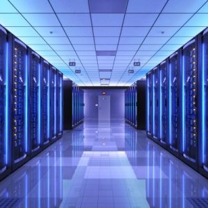 A spacious pathway lined with servers in racks on either side in the interior of a data center glowing with a blue hue.