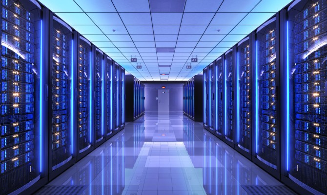 A spacious pathway lined with servers in racks on either side in the interior of a data center glowing with a blue hue.
