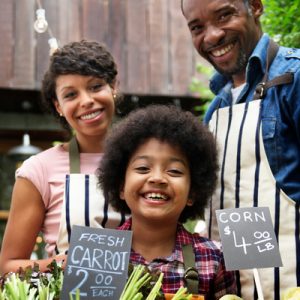 A smiling family standing together behind the array of fresh vegetables and produce they’re selling.