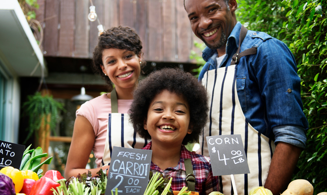 A smiling family standing together behind the array of fresh vegetables and produce they’re selling.