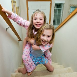 Two smiling girls in pajamas playing on the stairs. The older sibling is carrying the younger sibling on her back.