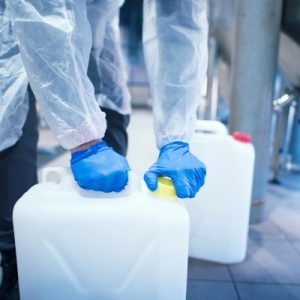 A person wearing a lab coat and protective rubber gloves twists the cap on a plastic can of chemicals in a lab.