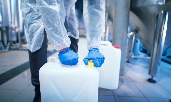 A person wearing a lab coat and protective rubber gloves twists the cap on a plastic can of chemicals in a lab.