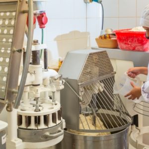 A young bakery worker pouring liquid into a professional kneading machine in an industrial bakery setting.