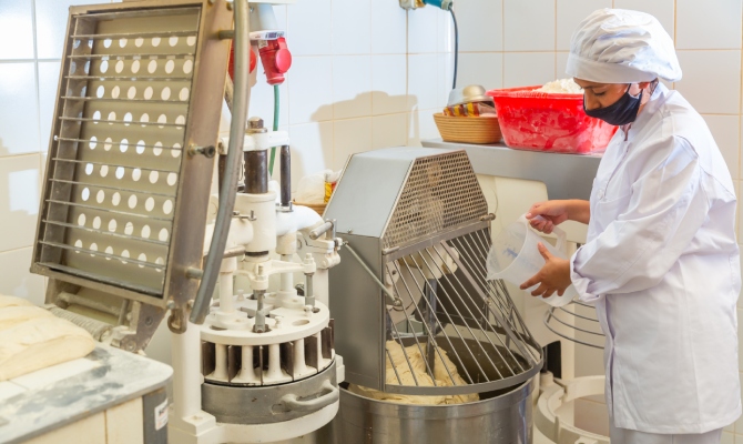 A young bakery worker pouring liquid into a professional kneading machine in an industrial bakery setting.