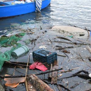 Plastic and microplastic waste in the water, with a fishing net and a wooden boat in the background.