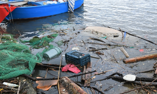 Plastic and microplastic waste in the water, with a fishing net and a wooden boat in the background.