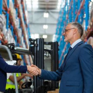 A man and a woman, both wearing navy suits, shaking hands while standing between pallet racks in a warehouse.
