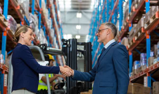 A man and a woman, both wearing navy suits, shaking hands while standing between pallet racks in a warehouse.