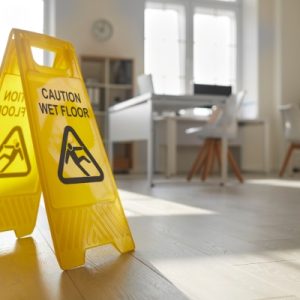 A bright yellow "Caution Wet Floor" sign sits on an office floor, with desks, chairs, and computers in the background.