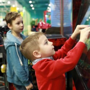 A child in a red shirt engaging with an interactive game on a touchscreen device with a child in green watching him.