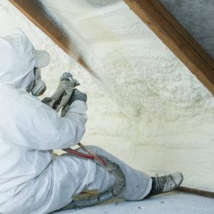 A person wearing a white protective suit and a white face respirator sits on the floor, installing spray foam insulation.