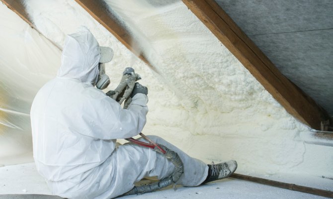 A person wearing a white protective suit and a white face respirator sits on the floor, installing spray foam insulation.