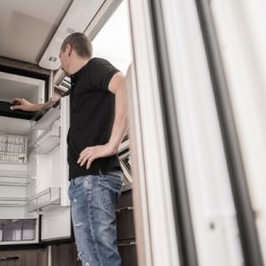 A man wearing a black t-shirt and jeans stands with his hand on his hip in front of an open black and silver fridge.