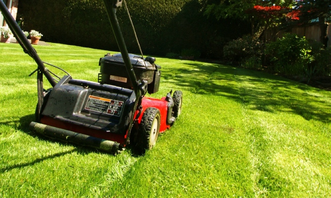 A lawn mower in action with someone pushing it off to the side. The lawn is green and healthy and the mower is red.