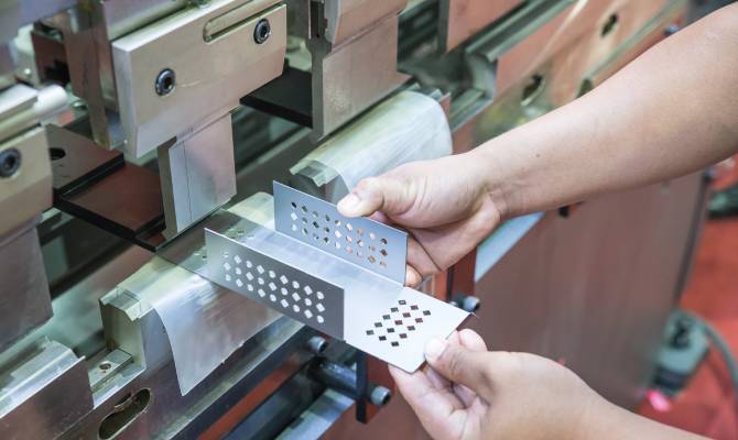 A manufacturer placing a metal component into a metal stamping machine. The metal part has punched holes throughout.