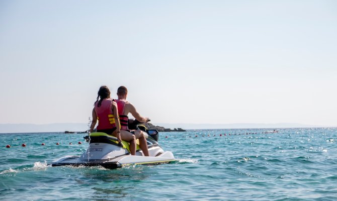 A couple rides on a jet ski on the ocean. The waves go on for a long time while the sky is clear and free of clouds.