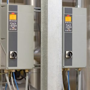 Two variable frequency drives with several buttons and cords hanging from their base in an industrial setting.