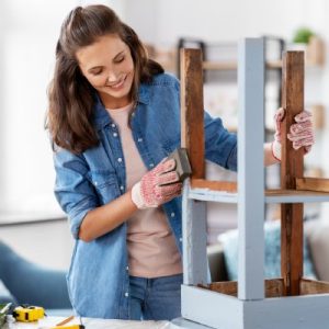 A woman uses sanding paper to sand down the sides of an upside-down stool. The stool is partially painted light blue.