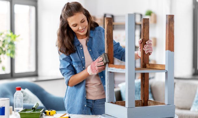A woman uses sanding paper to sand down the sides of an upside-down stool. The stool is partially painted light blue.