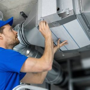A technician wearing a blue shirt and hat works on a ventilation system air duct on the ceiling in a room.