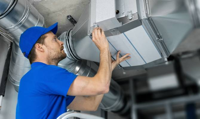 A technician wearing a blue shirt and hat works on a ventilation system air duct on the ceiling in a room.