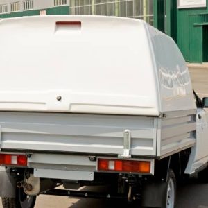 A white pickup truck with a large white truck topper installed over the bed. The truck is parked near a large green building.