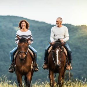 A man and a woman ride horses together in a field with mountains in the background. Both parties have smiles on their faces.