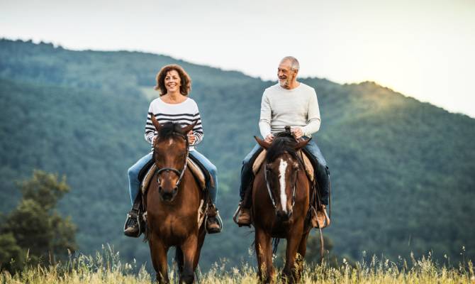 A man and a woman ride horses together in a field with mountains in the background. Both parties have smiles on their faces.