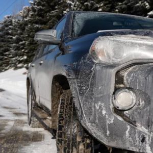 The front of an SUV is covered in white marks from moisture and road salt. Snow and trees are seen on the side of the road.