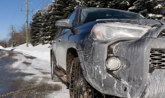 The front of an SUV is covered in white marks from moisture and road salt. Snow and trees are seen on the side of the road.