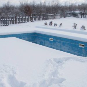A large backyard swimming pool surrounded by a foot of snow. The cover is off the pool and the water is blue.
