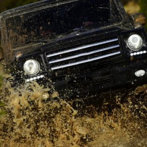 A black off-roading vehicle drives through muddy water. The tires splash mud onto the windshield.