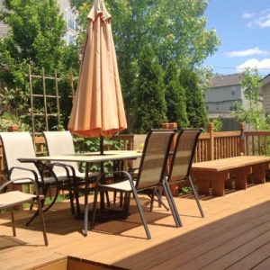 A home’s exterior deck with wood flooring and posts, an umbrella table with chairs, and trellises and plants.