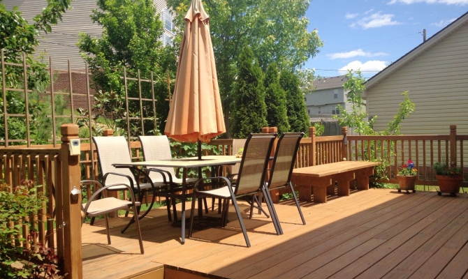 A home’s exterior deck with wood flooring and posts, an umbrella table with chairs, and trellises and plants.