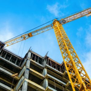 A view from the ground focuses on a tall yellow crane next to a building under construction. A blue sky is overhead.
