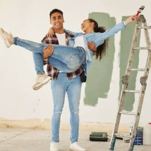 A young, happy couple standing in their remodeled home. They're next to a ladder with green paint swatches on the wall.