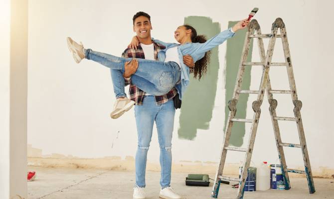 A young, happy couple standing in their remodeled home. They're next to a ladder with green paint swatches on the wall.