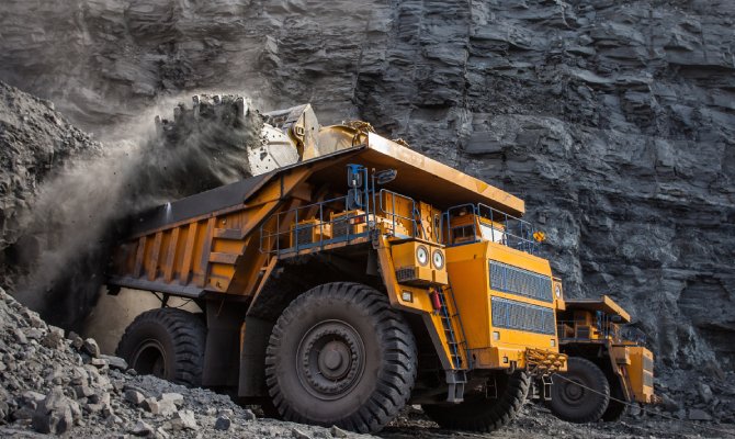  A large yellow mining truck in a quarry is loading a massive amount of coal and coal dust into the rear compartment.