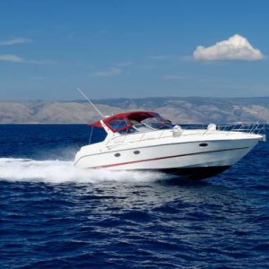 A white motorboat with a red cover and red stripe speeding through the water on a blue lake with hills and a partly cloudy sky in the background.