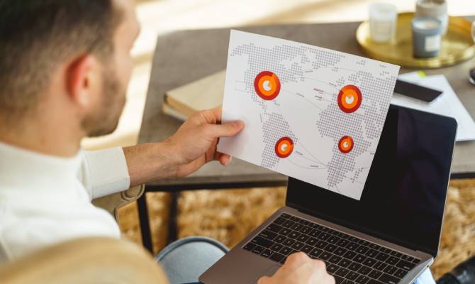 A man sits at a desk with a laptop on his lap and a piece of paper with a flight plan in his left hand.