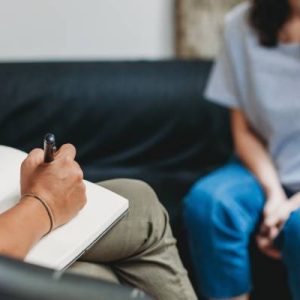 A therapist writing on a white notepad with a pen while talking with a patient sitting across from her on a dark couch.