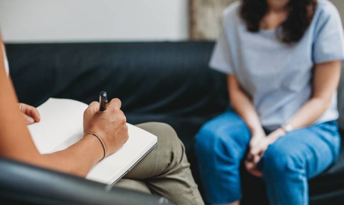 A therapist writing on a white notepad with a pen while talking with a patient sitting across from her on a dark couch.